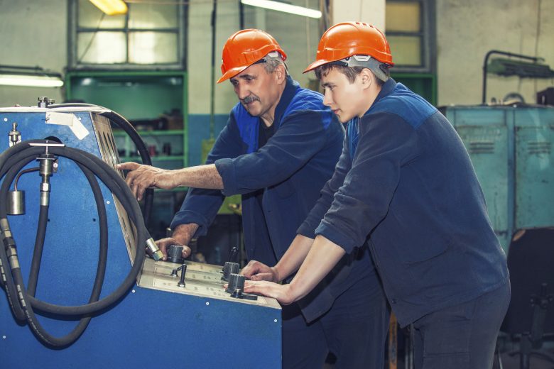 An older worker points out something on a machine for a younger worker to see