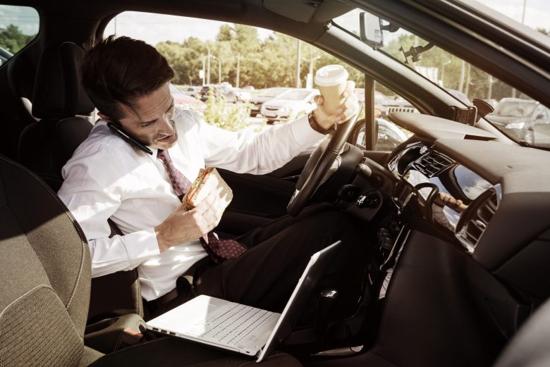 Distracted working businessman eating while on his phone and laptop driving his car