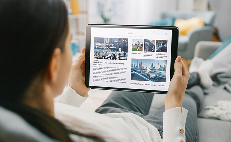 Over the Shoulder Shot of a Young Woman at Home Lying on a Couch and Using Tablet Computer in Horizontal Landscape Mode for Reading News about Technology. Girl Using Touchscreen Device.