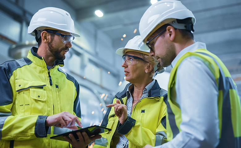 Three Heavy Industry Engineers Stand in Pipe Manufacturing Factory, Use Digital Tablet Computer, Have Discussion. Large Pipe Assembled. Design and Construction of Oil, Gas and Fuels Transport Pipeline